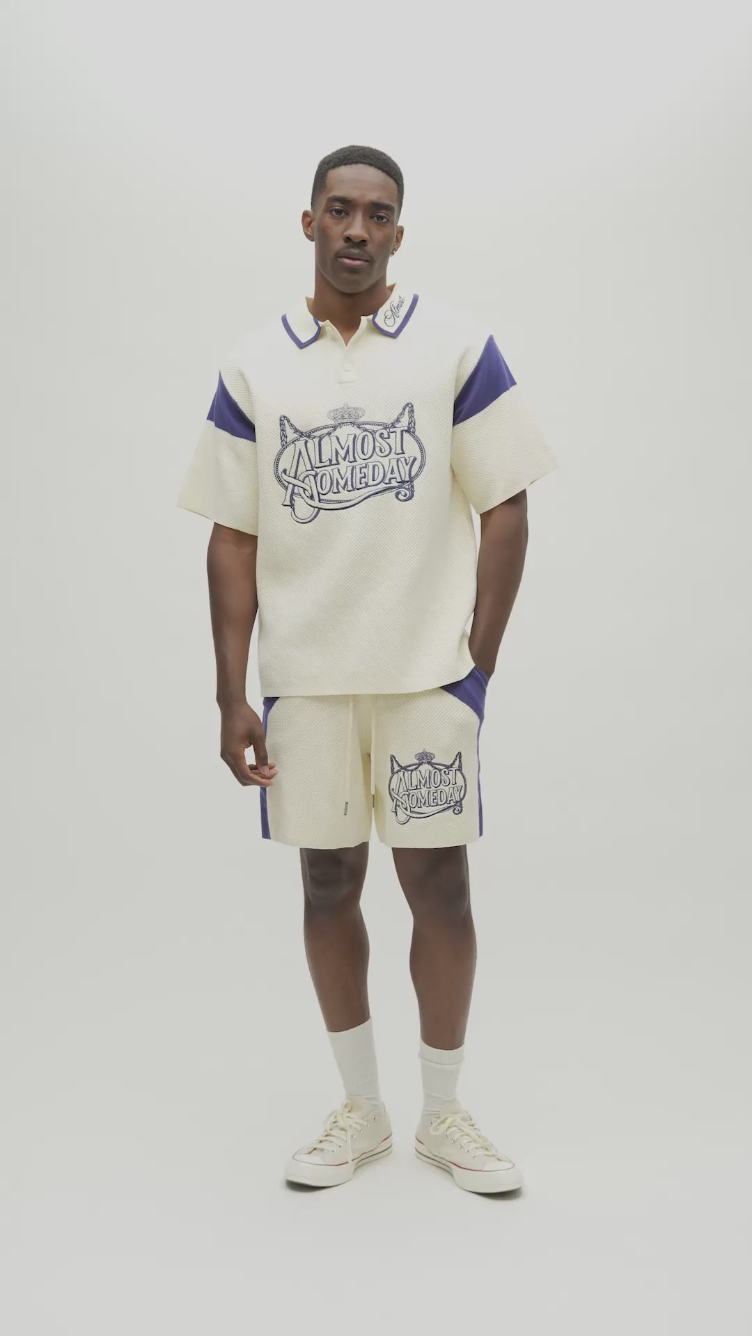 A man modeling a cream-colored short-sleeve polo shirt with blue accents and a graphic print on the front, paired with matching cream pants with blue side panels, against a plain light gray background.