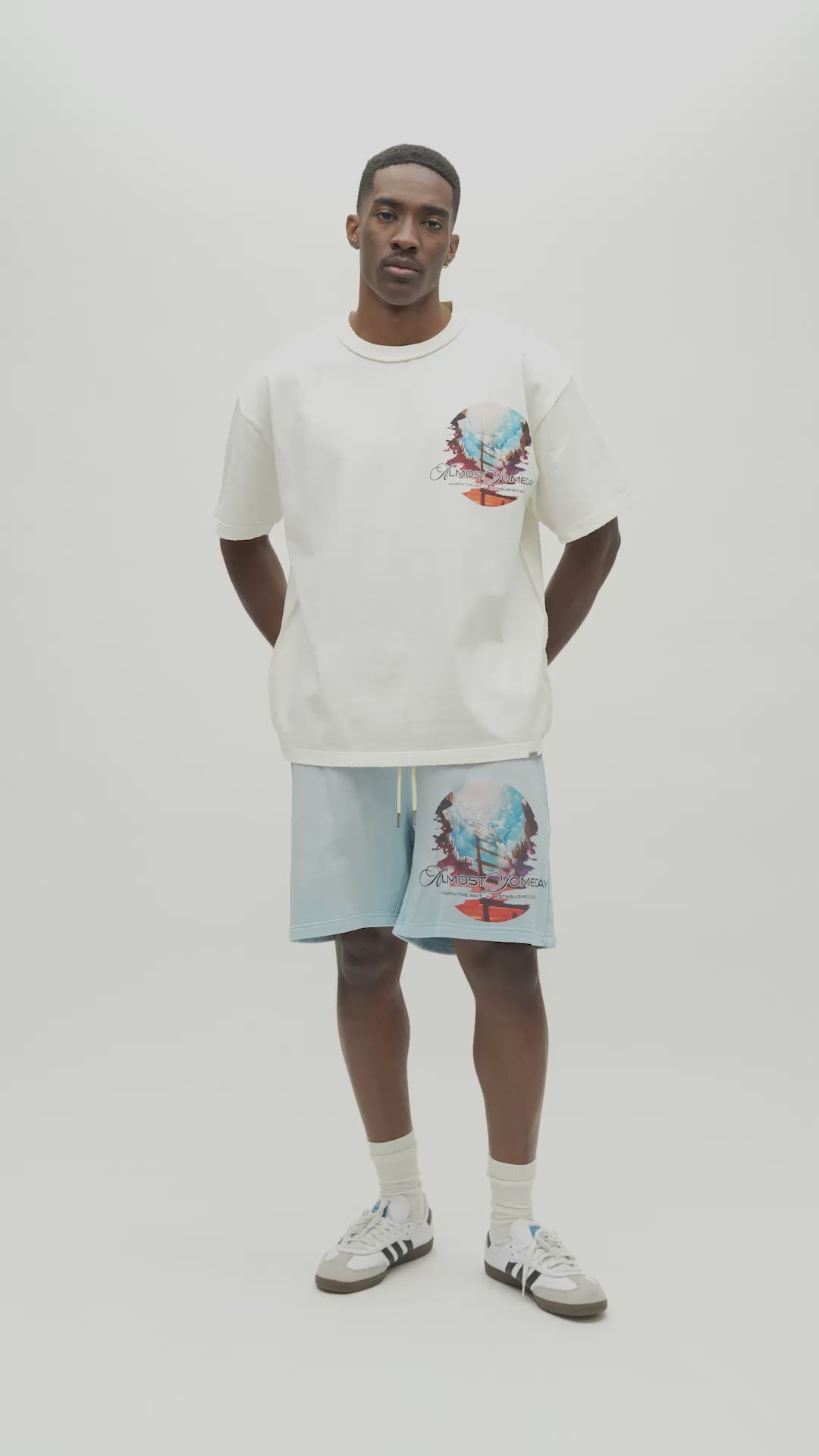 A man wearing a white Almost Someday graphic t-shirt with a large colorful print of a sailing ship and text, paired with light blue sweatshorts against a plain white background.