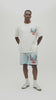 A man wearing a white Almost Someday graphic t-shirt with a large colorful print of a sailing ship and text, paired with light blue sweatshorts against a plain white background.