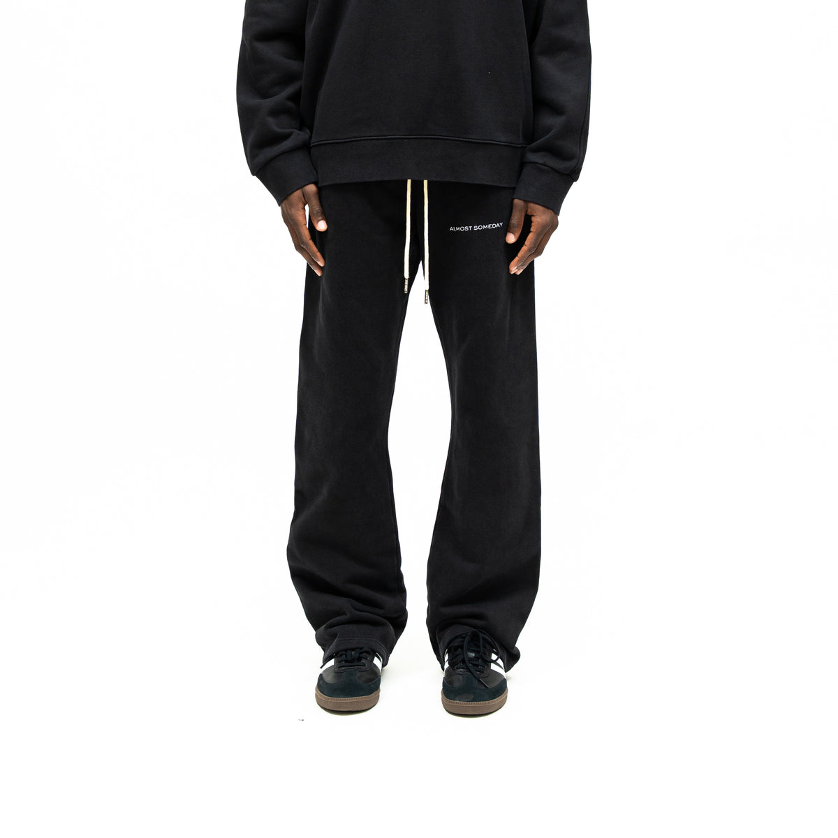 Smoke Flare Bottoms – Black – Front view on model styled with black crewneck, showing cream drawstrings and white thigh lettering